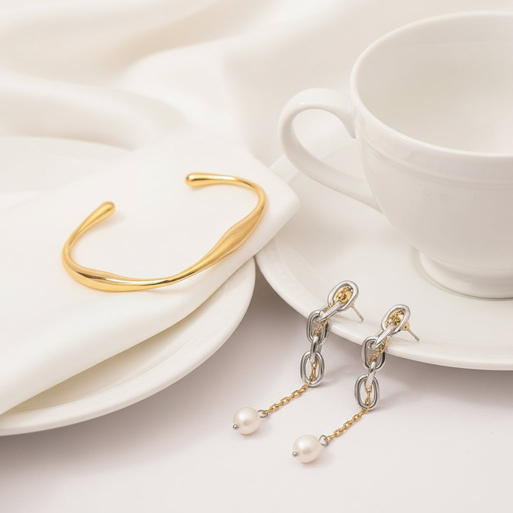 Gold bracelet and silver earrings with pearl drops on a white surface with a cup and saucer.