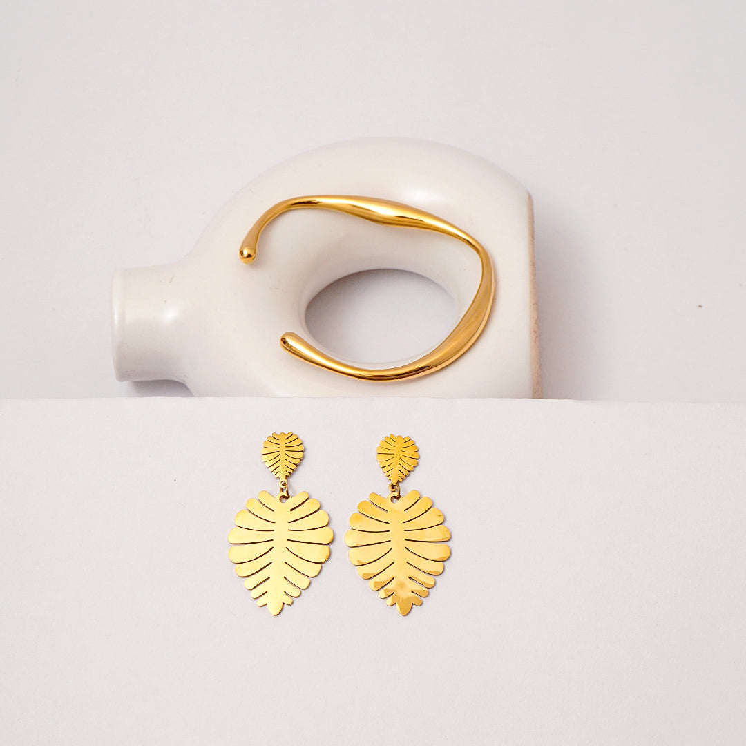 Gold leaf-shaped earrings on a white surface with a gold ring in the background