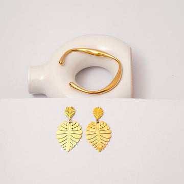 Gold leaf-shaped earrings on a white surface with a gold ring in the background