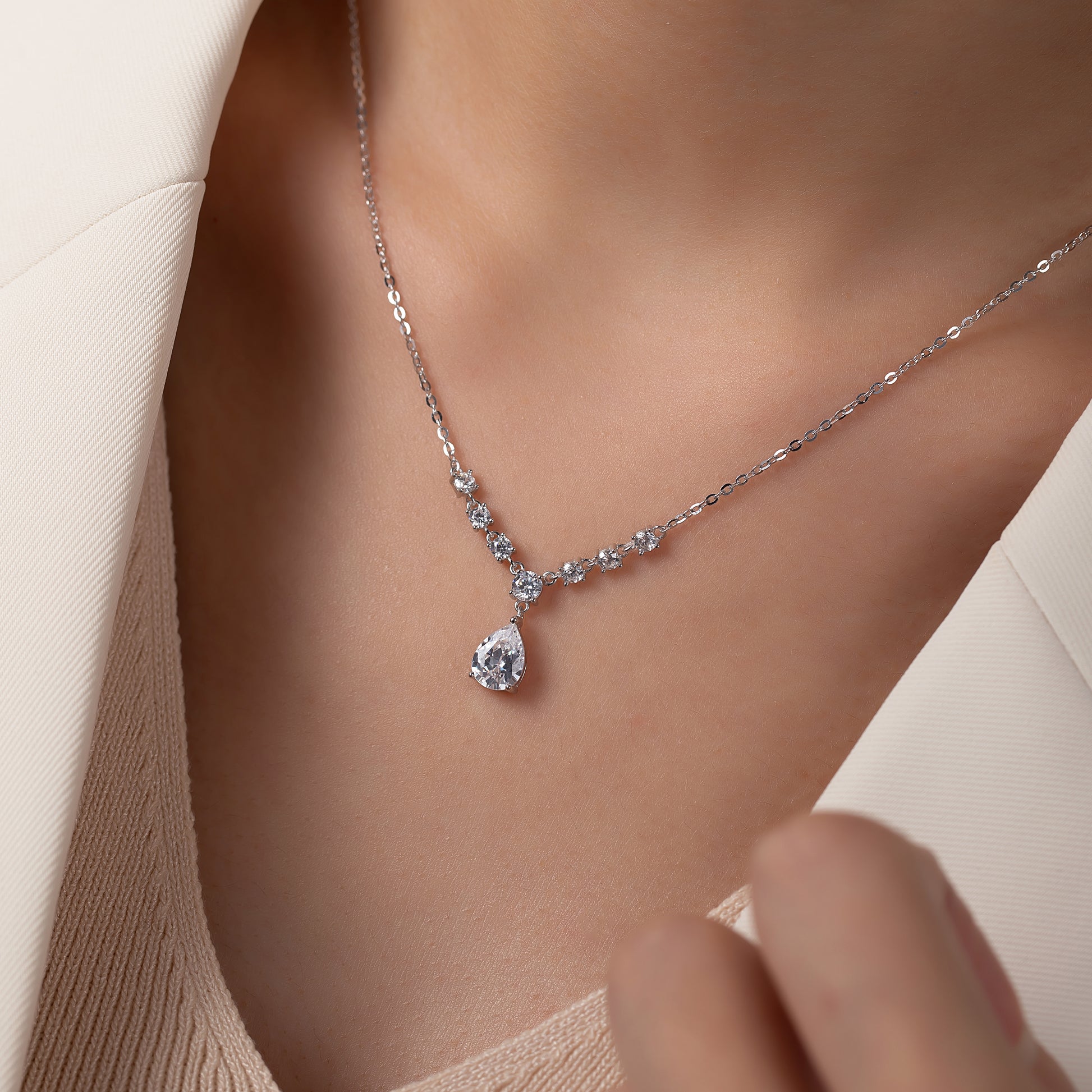 Delicate silver necklace with a teardrop-shaped pendant on a person wearing a beige outfit.