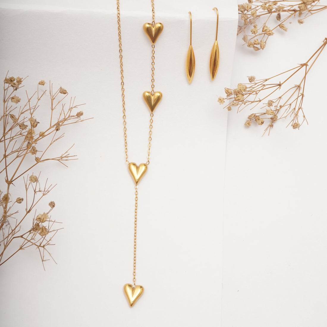 Gold necklace with heart-shaped pendants on a white background with dried flowers.