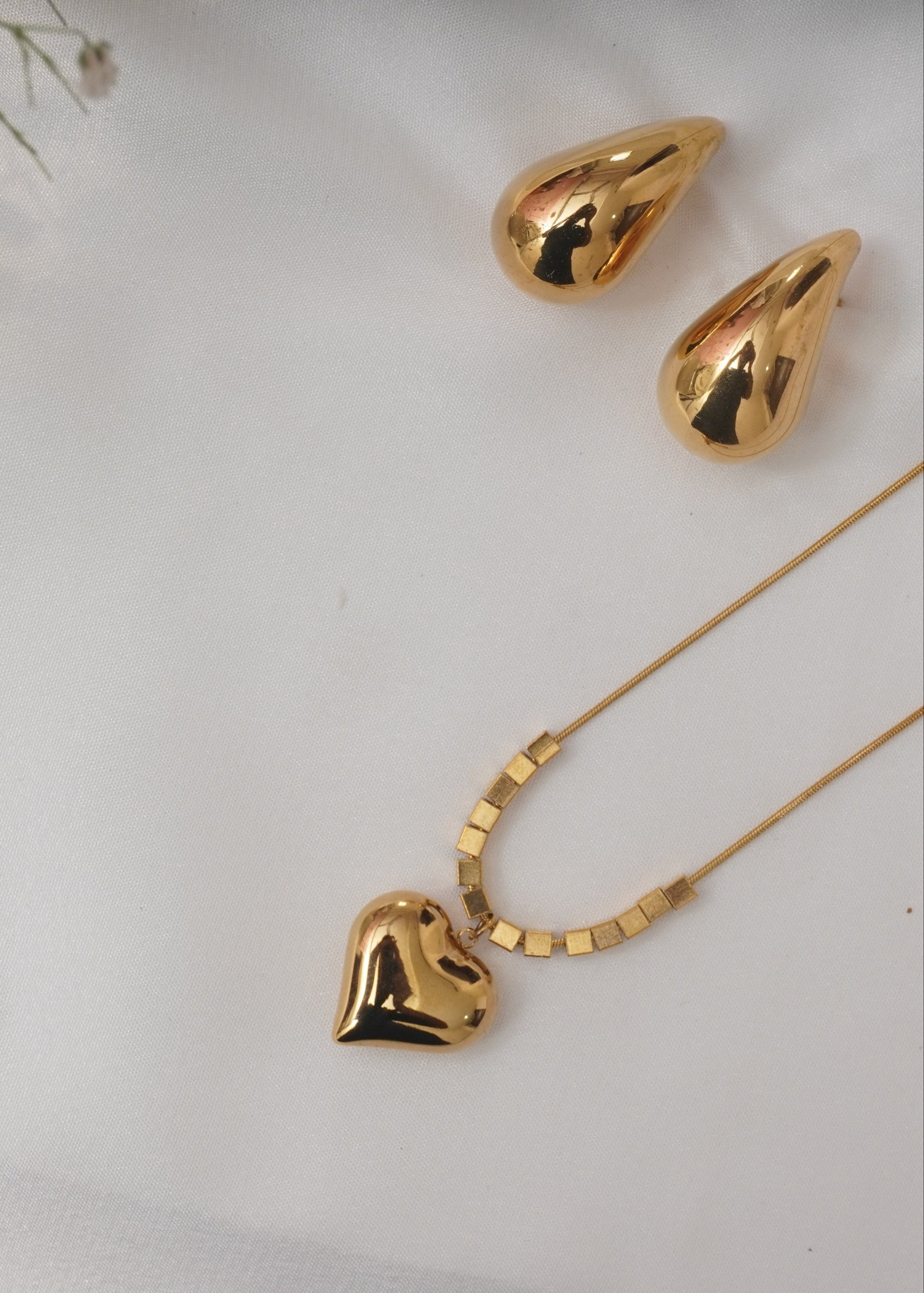 Gold heart-shaped necklace and teardrop-shaped earrings on a white background with small flowers.